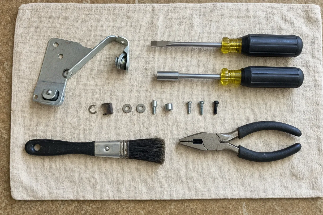 Appliance door hinge and its roller laid out on canvas beside a nut driver, a flat driver, a dust brush and pliers